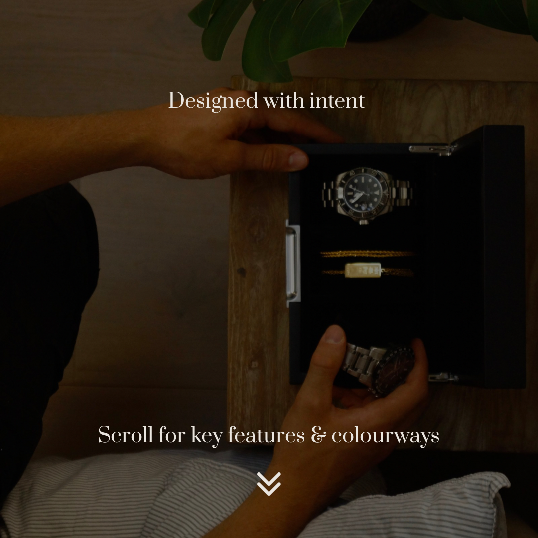 Bird’s-eye lifestyle action image of the LDN III men’s 3-slot black watch box by Transform, shown open on a bedside table holding two watches and two necklaces. A man’s arms are visible as he removes one of the watches. Text overlay reads ‘Designed with intent’ at the top and ‘Scroll for key features and colourways’ underneath.