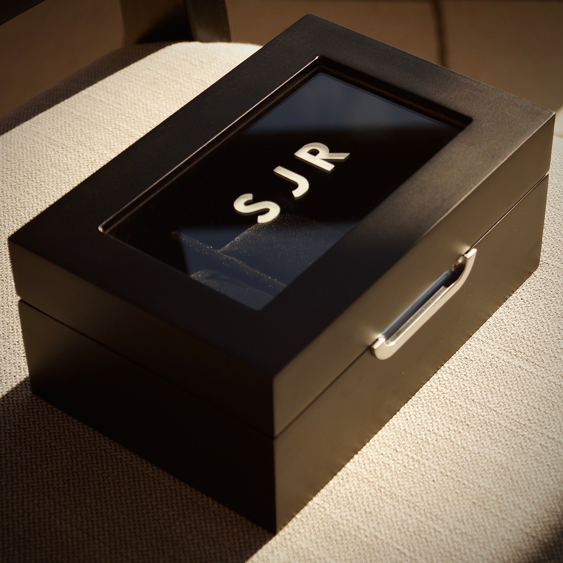 Bird’s-eye angled lifestyle image of the LDN III men’s 3-slot black watch box by Transform, shown closed and placed on a chair, featuring personalisation with engraved text on the glass lid.
