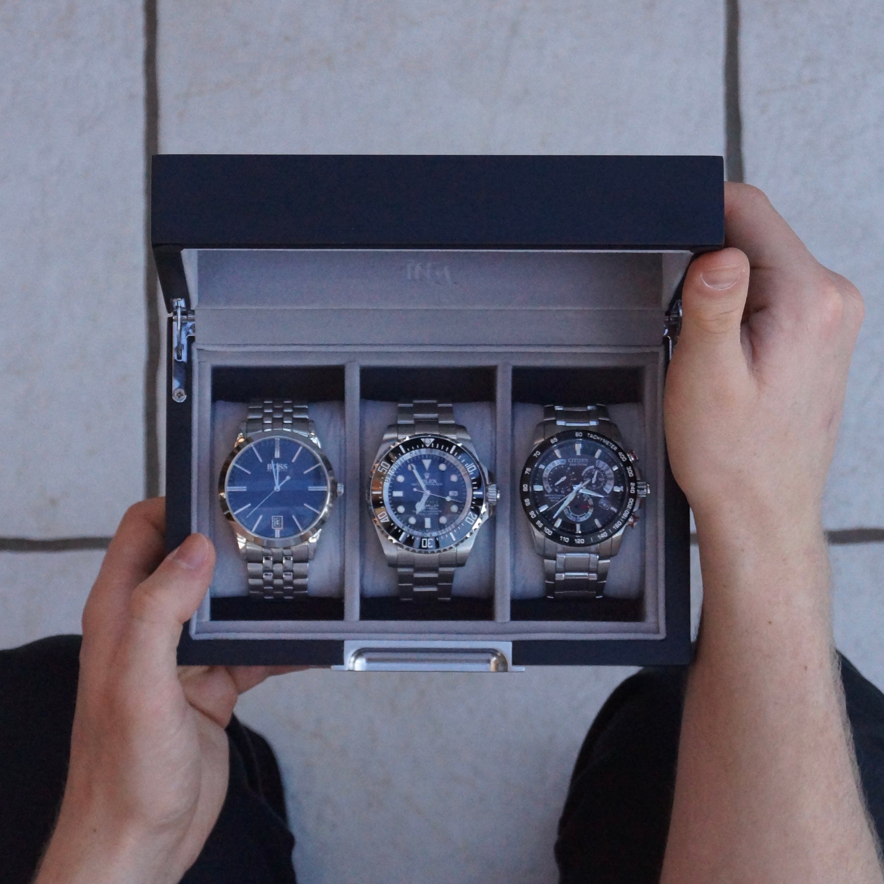 Three wristwatches are displayed in a grey multi-watch box held open by two hands over a light tiled floor.