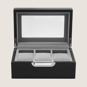 A black wooden watch box with an open hinged lid featuring a clear glass window and three interior watch slots, shown on a light beige background.
