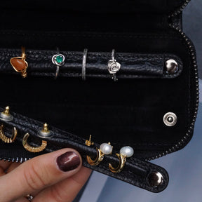 A close-up of a black zippered jewellery travel case being held open by a hand. The pouch has two horizontal slots holding rings and two lower slots holding gold hoop earrings and pearl studs.