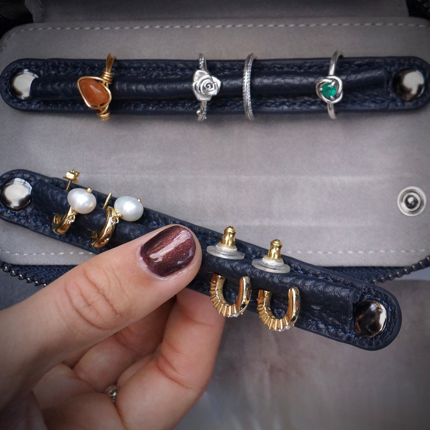 A close-up of a hand holding a dark blue leather jewellery travel case with multiple rings and earrings displayed on its slots, set against a grey textured background.
