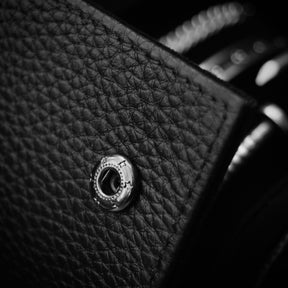 Close-up of a black pebbled leather watch roll with a silver-tone metal snap closure, photographed in dramatic black-and-white lighting.