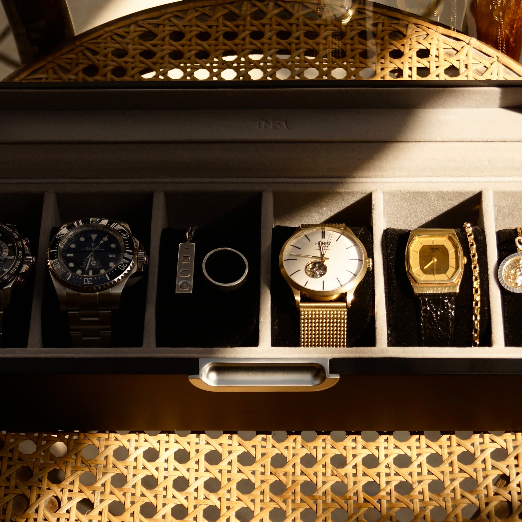 Close-up, front-angled lifestyle view of the grey LDN VI men’s 6-slot watch box by Transform, shown on a chair and filled with watches, necklaces and rings.