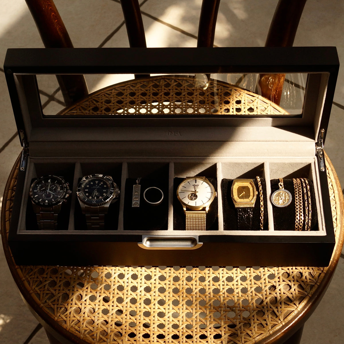 Front-angled lifestyle image of the grey LDN VI men’s 6-slot watch box by Transform, placed on a chair and filled with watches, necklaces and rings.