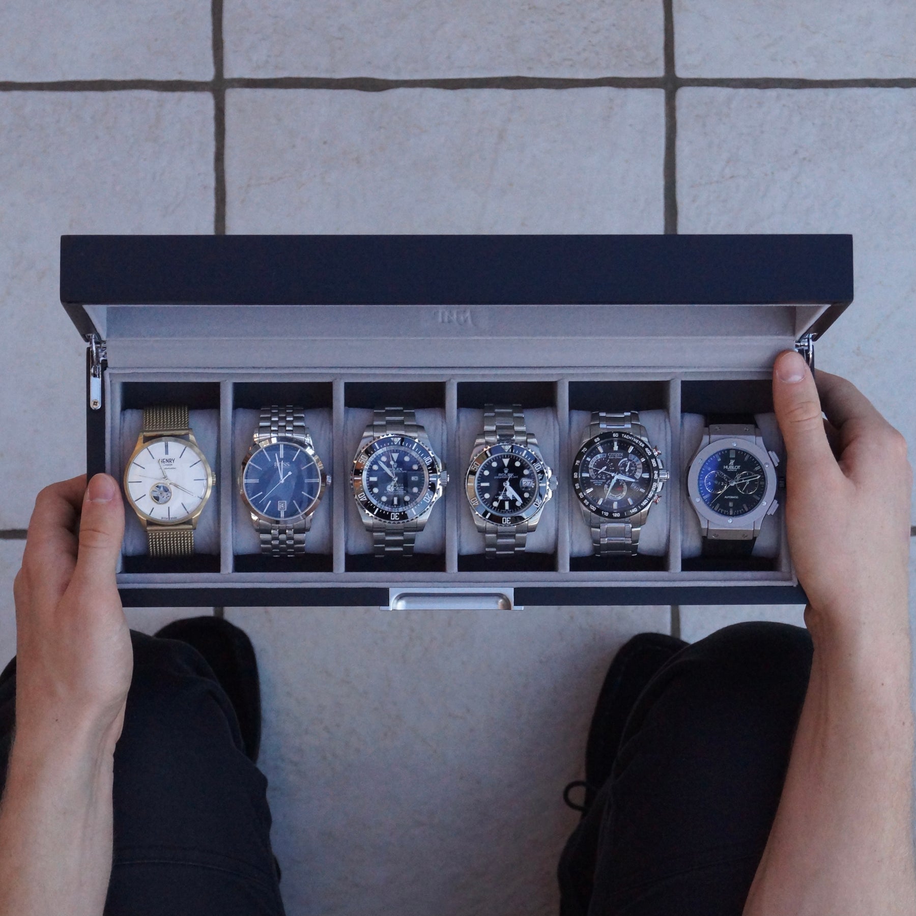 A top-down view of a person holding open a black watch display box containing six wristwatches, set on a light tiled floor.