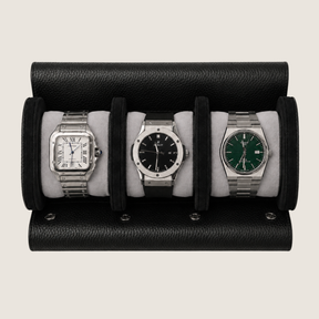 A black textured leather watch roll holding three wristwatches on light grey cushions, photographed against a plain light beige background.