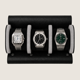 A black pebbled leather watch roll holding three wristwatches side by side on a light beige background.