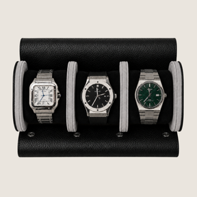 A black pebbled leather watch roll holding three wristwatches side by side on a light beige background.